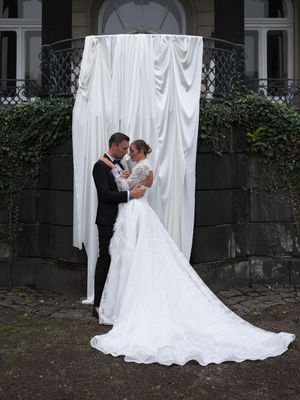 A romantic wedding couple poses against white fabric draping in a garden setting, creating an intimate portrait series.