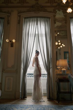 A silhouetted figure in a wedding dress stands gracefully by sunlit windows in an elegant room with flowing curtains.