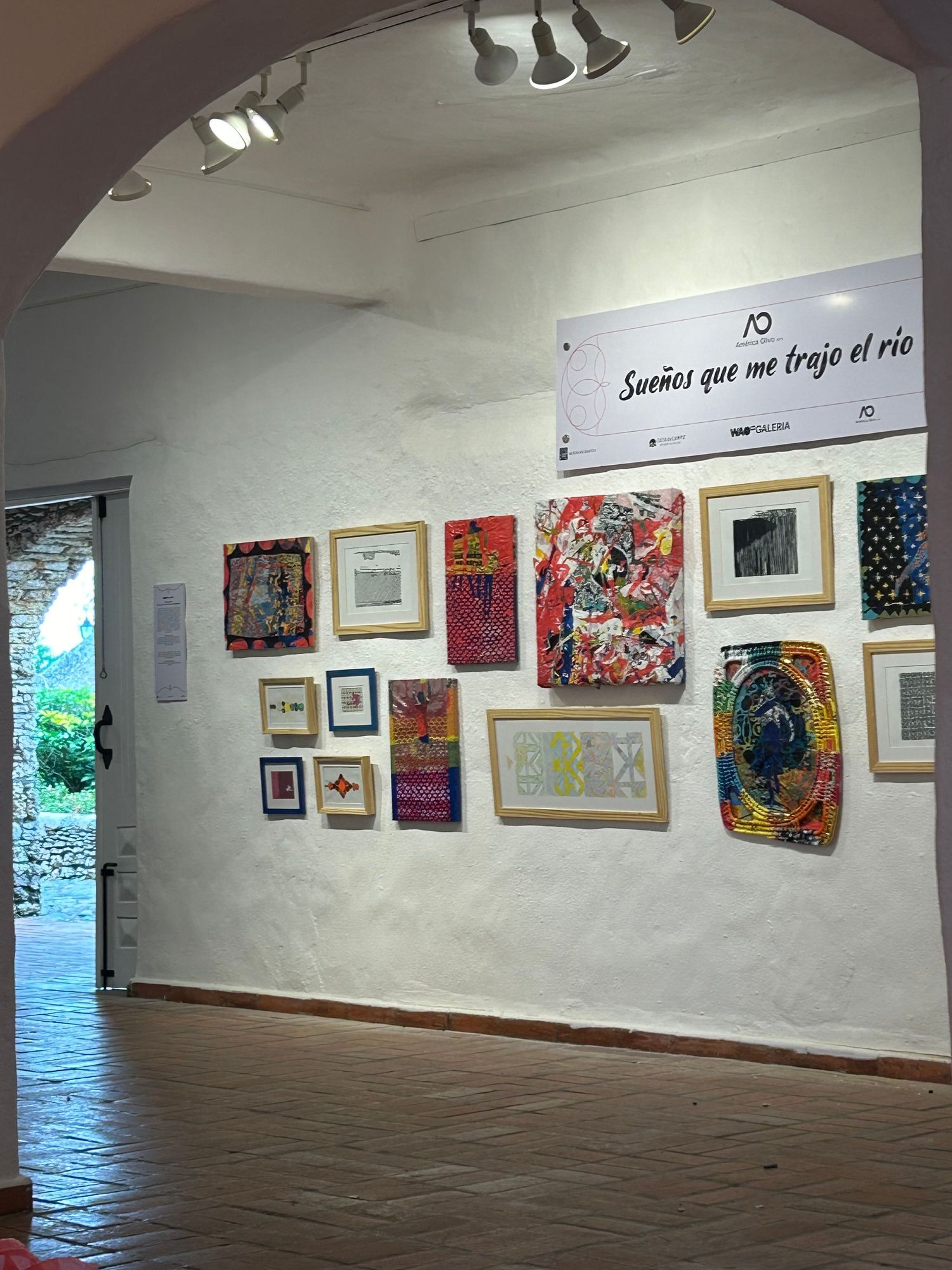 Art gallery wall displaying multiple colorful framed artworks and a text sign in a white-walled space.