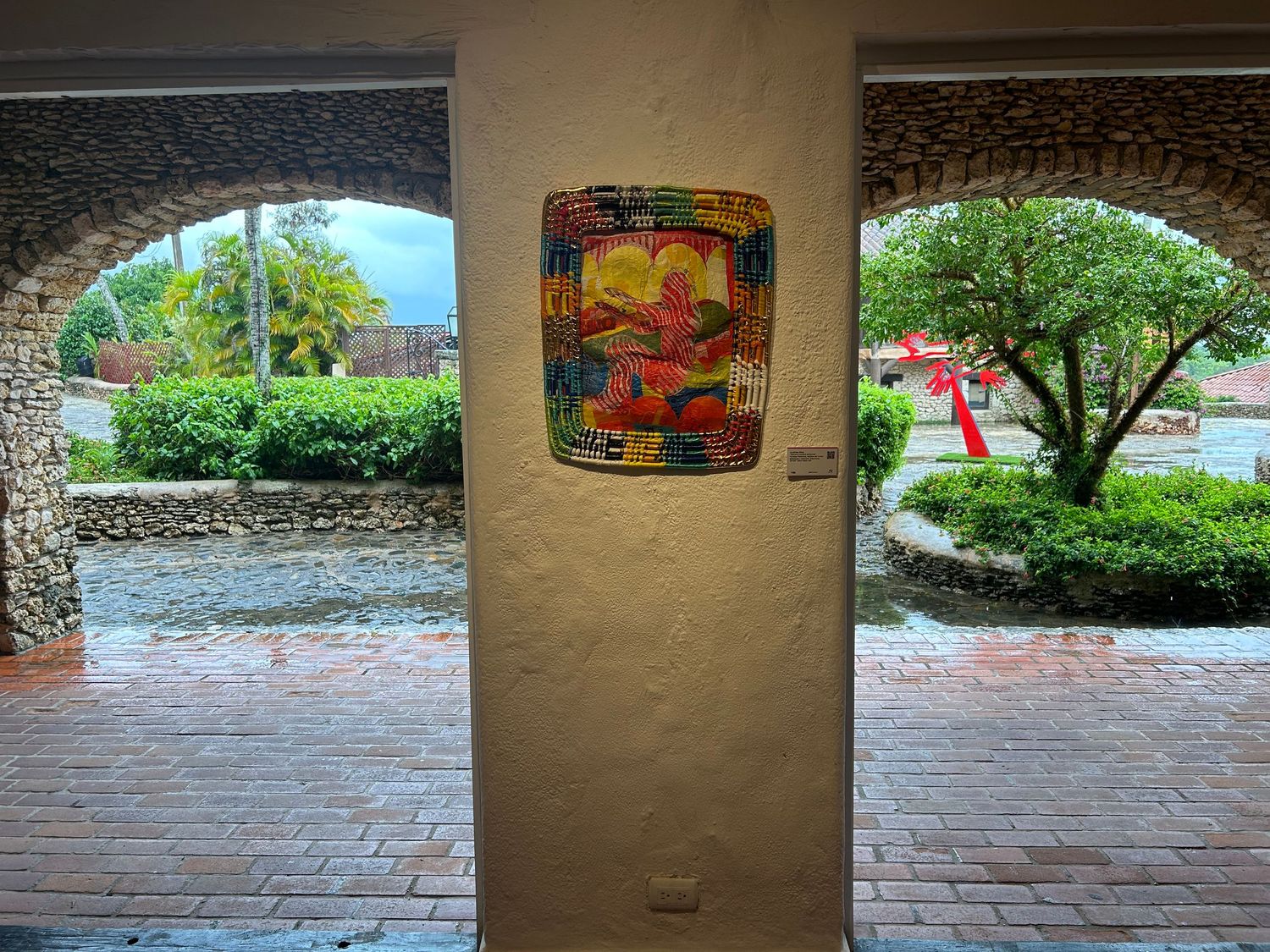 Colorful art installation displayed on pillar between arched walkways leading to outdoor garden view.