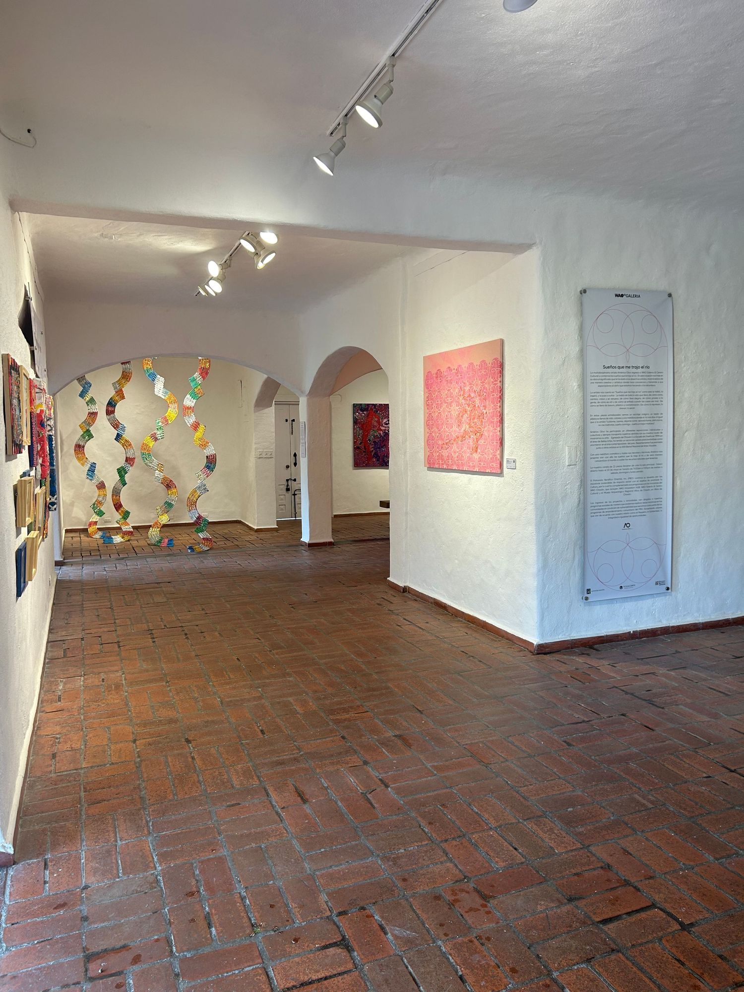 Art gallery hallway featuring colorful suspended installation and pink artwork on white walls.