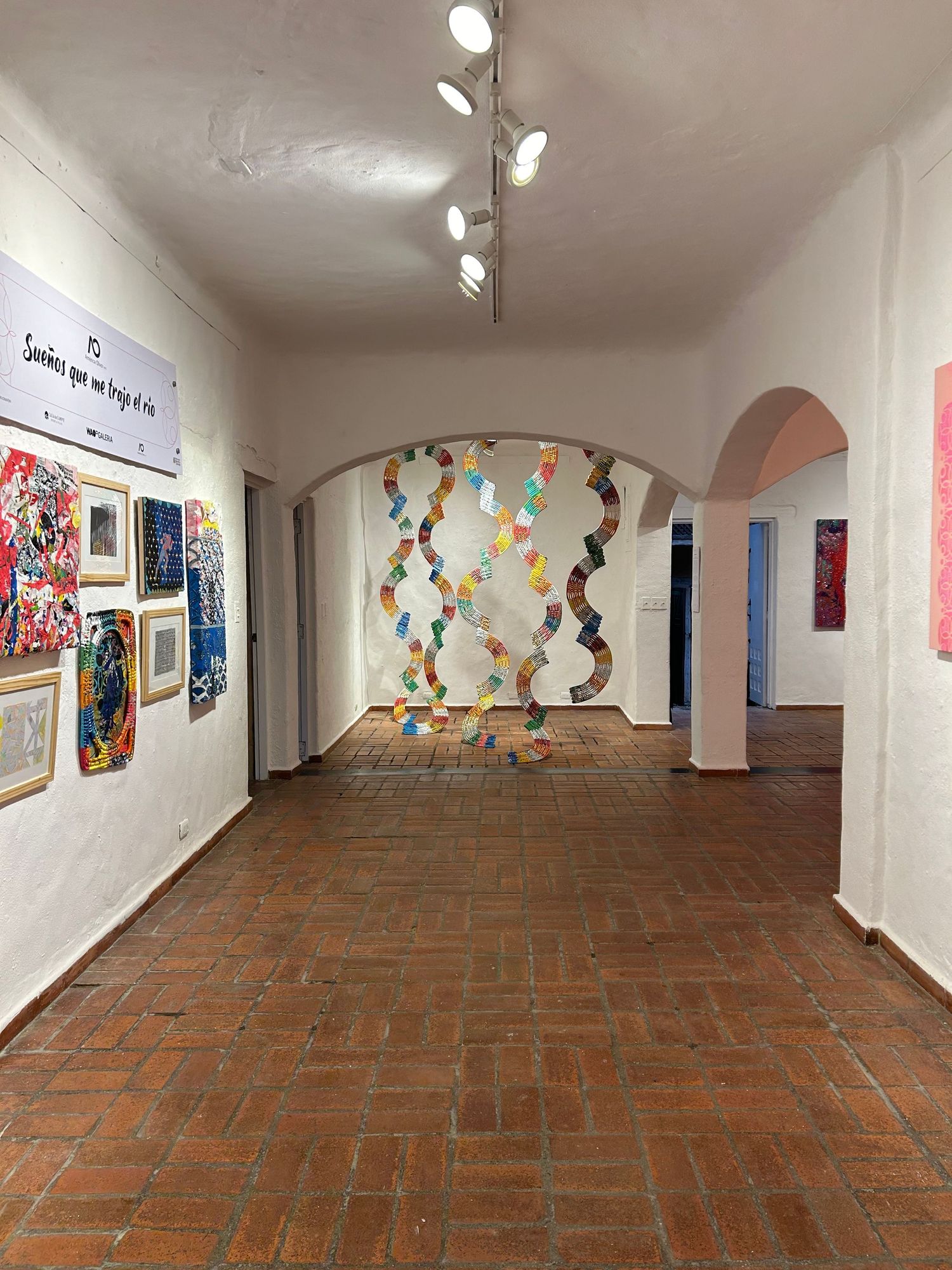 Gallery corridor with hanging geometric installation and diverse artworks mounted on white walls.