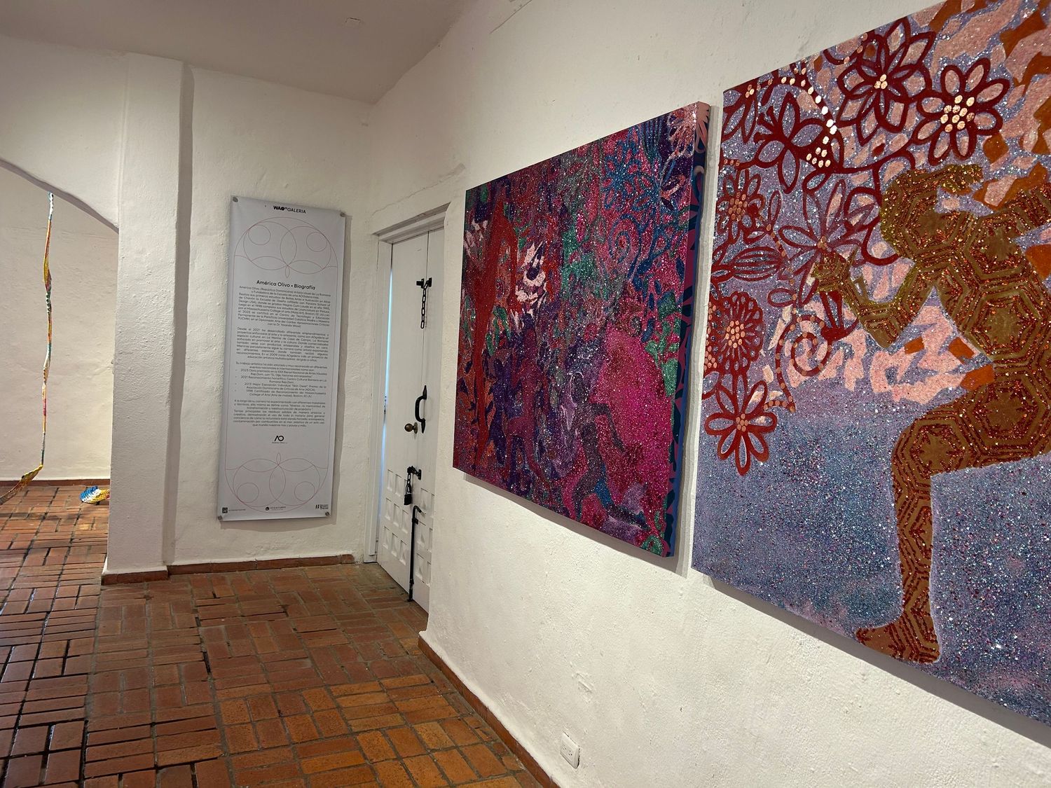 Gallery corridor with pink and purple abstract artwork displayed on white walls above brick flooring.