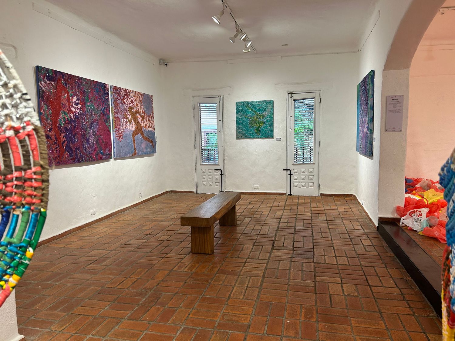 Gallery space with wooden bench, colorful art pieces, and vibrant decorations on brick floor.