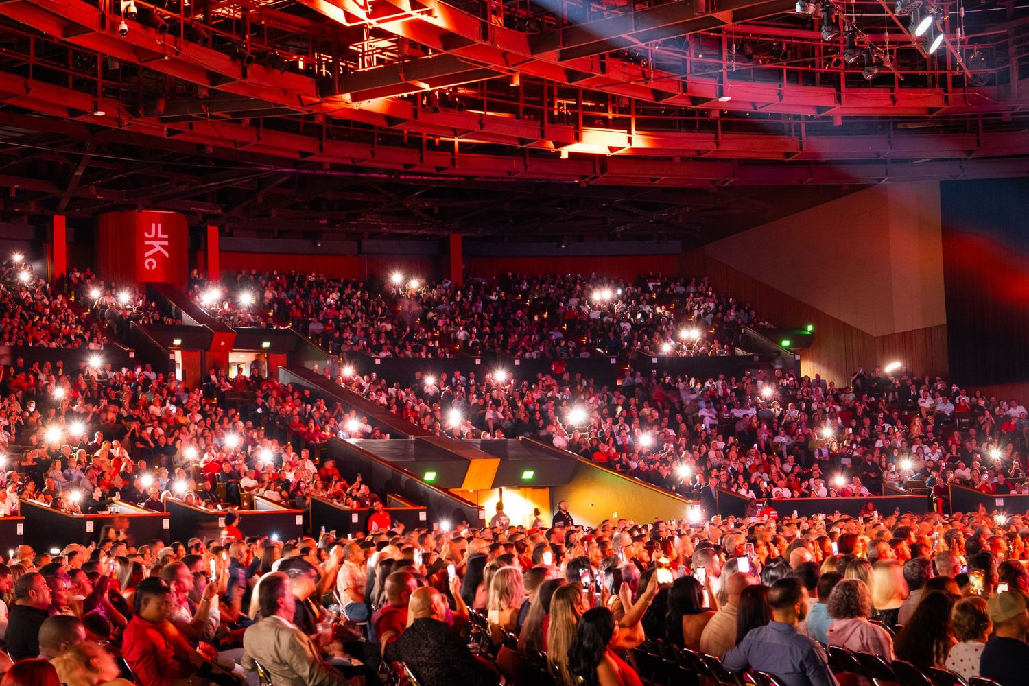 A massive crowd fills an arena bathed in red light, with audience members holding up glowing phone lights during a live event.