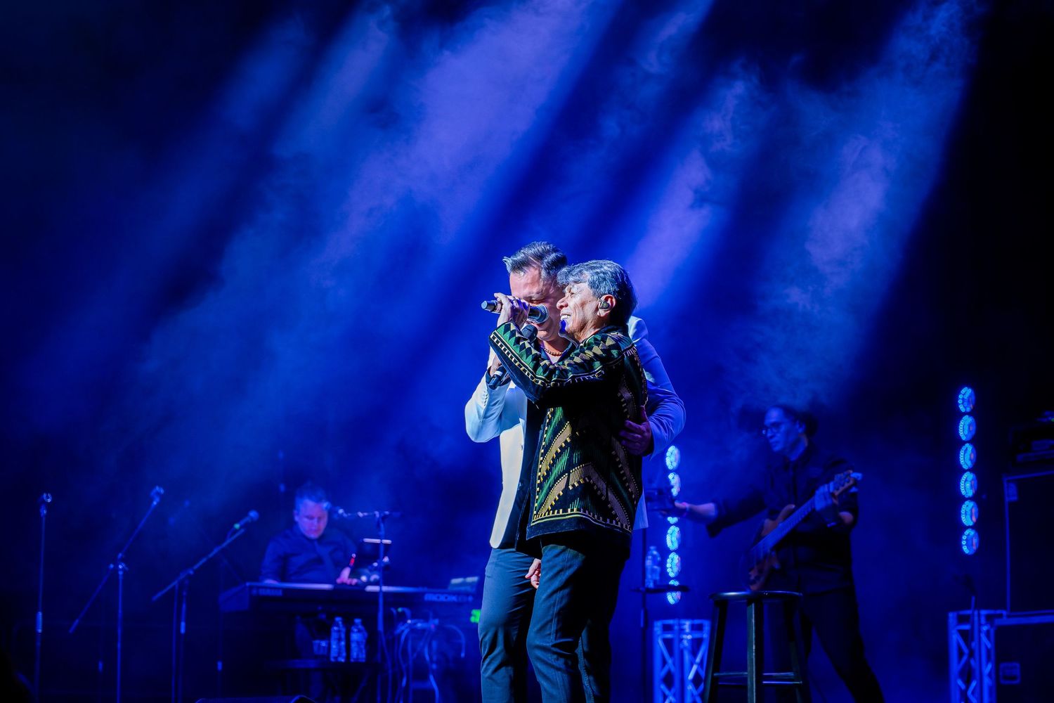 Two performers embrace on stage under dramatic blue lighting during a live concert performance.