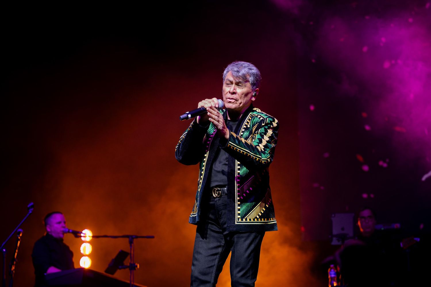 Male singer performing on stage in embroidered jacket, holding microphone under colorful concert lights.