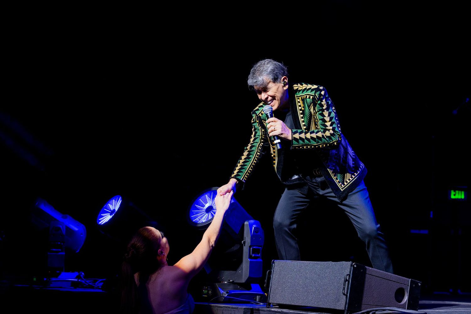 Elvis impersonator in sequined jacket hands beads to fan during energetic live stage performance under blue spotlight.
