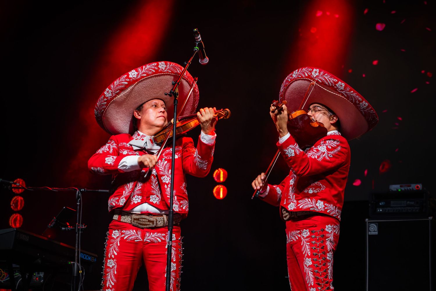 Two mariachi musicians in red charro suits and sombreros playing violins on a dimly lit stage.