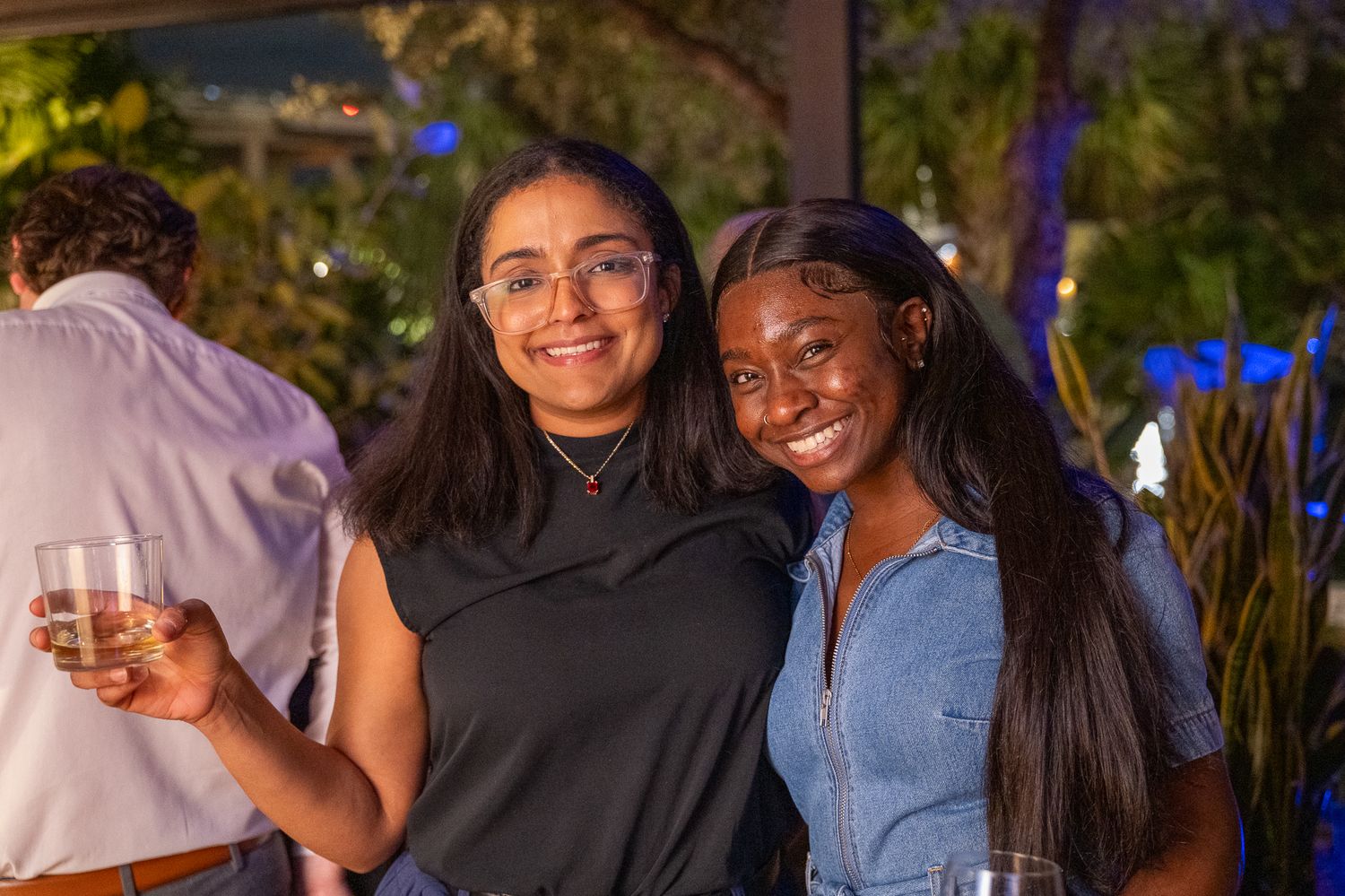 Two smiling women posing together at an outdoor evening party, holding drinks amid blue-lit tropical decor.