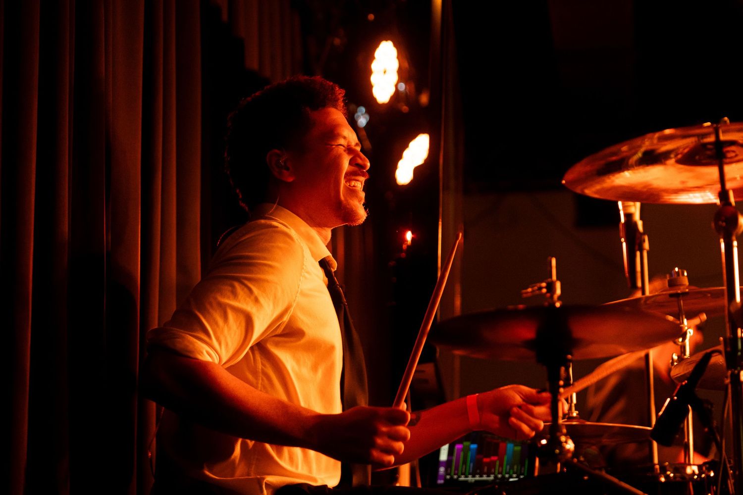 A drummer performs energetically on stage in moody lighting during a live music performance.