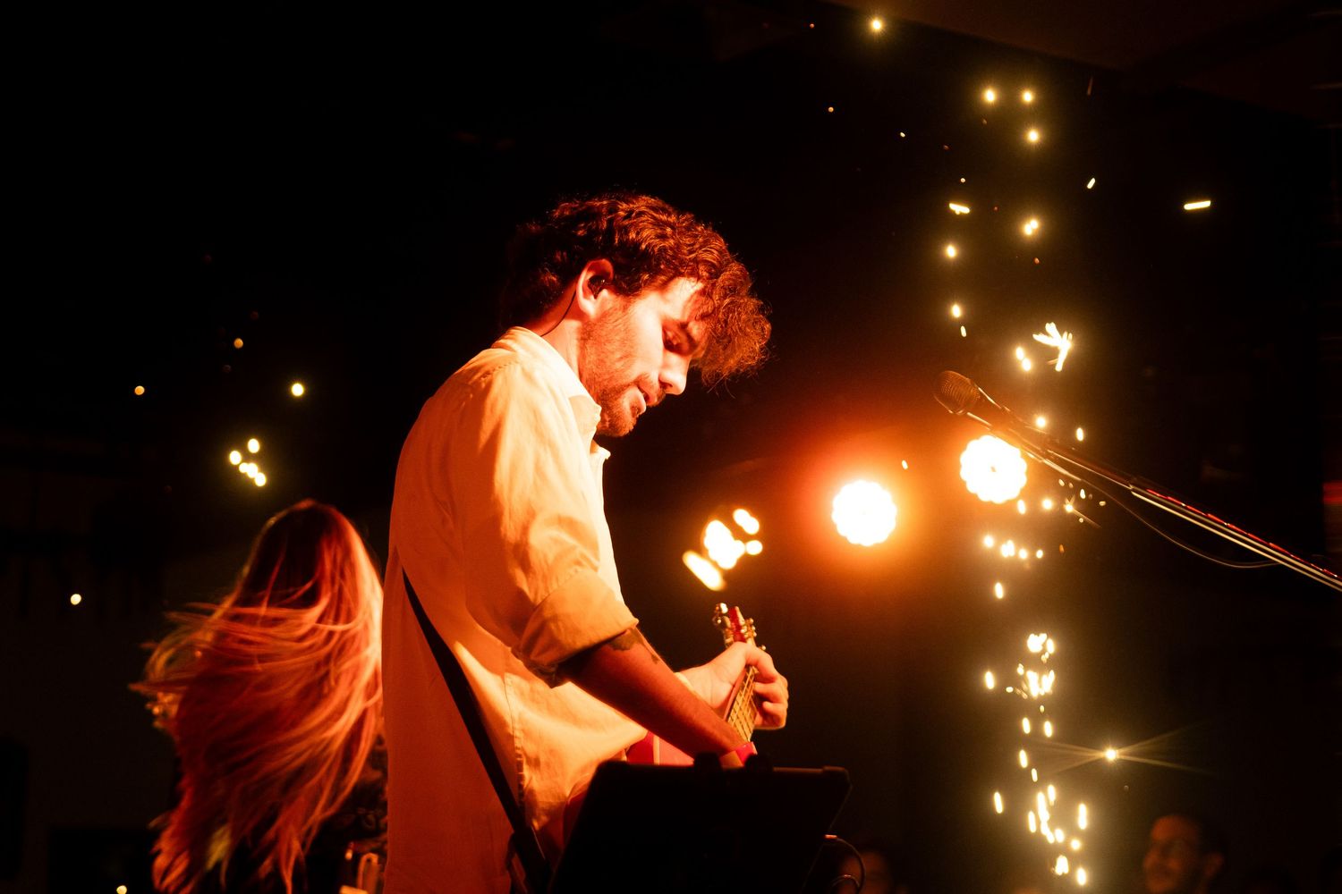 A musician performs on stage with a guitar against a backdrop of warm stage lights and glowing sparkles.