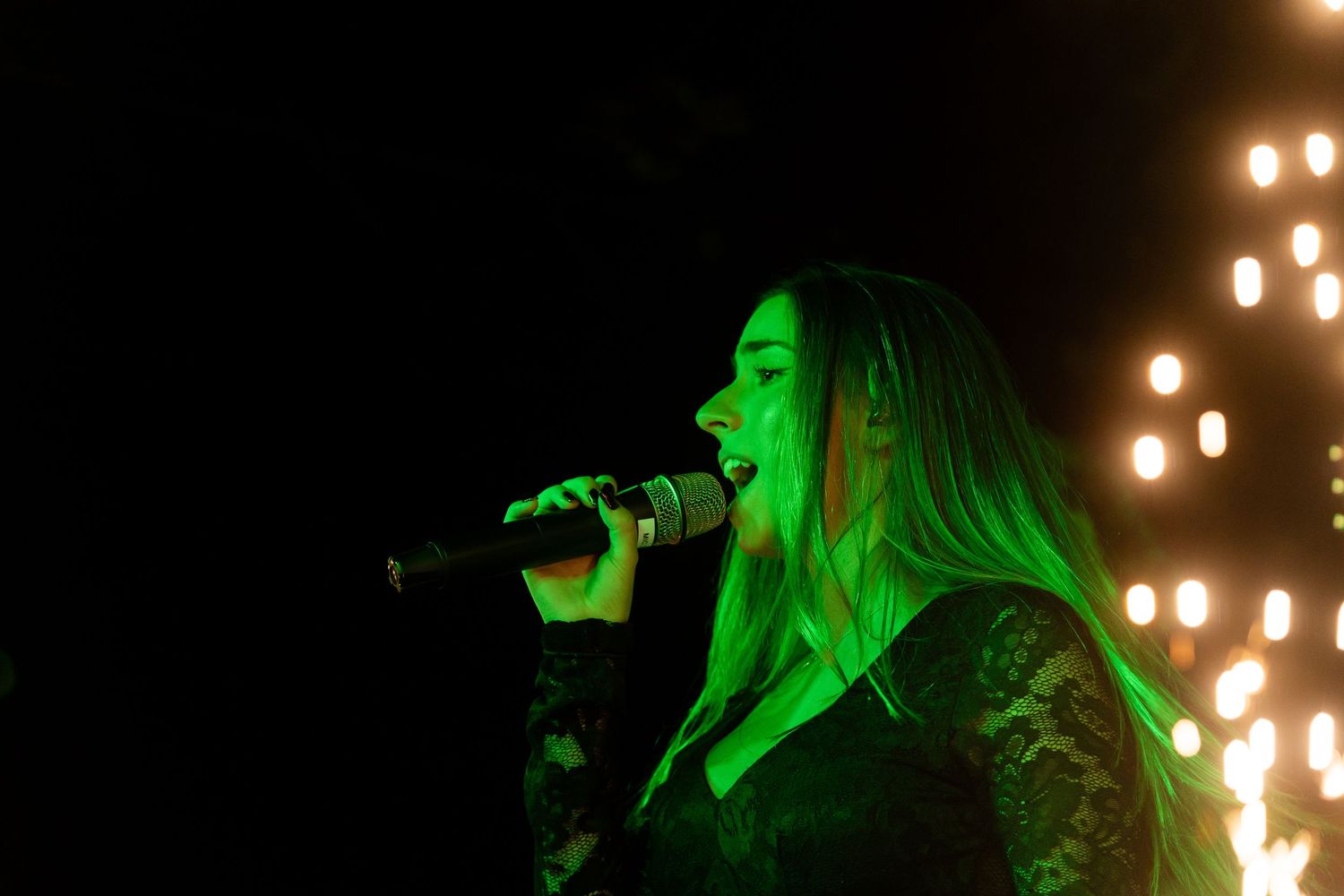 A performer sings passionately on stage illuminated by dramatic green lighting and glowing lights in the background.