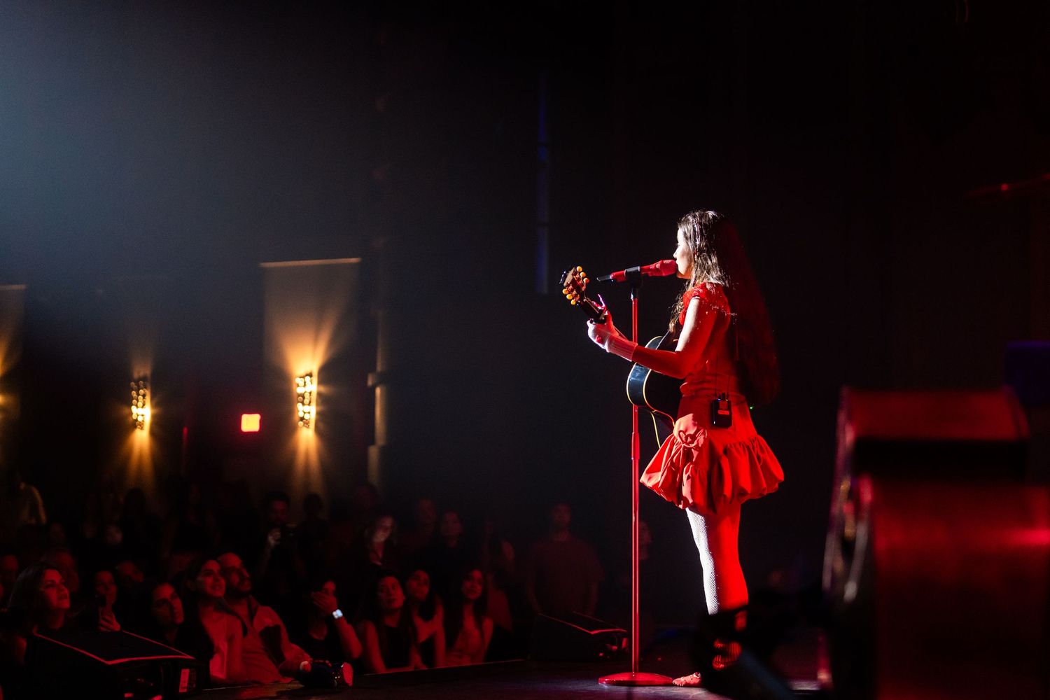 Joaquina in a red dress plays guitar on stage with dramatic lighting and a silhouetted audience in the background.