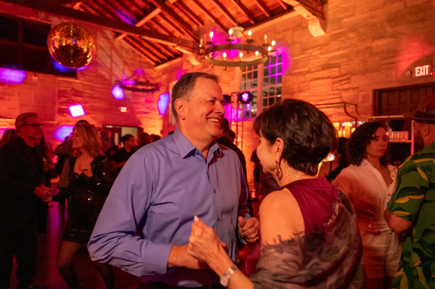Couple dancing joyfully at a lively party venue with warm pink lighting and disco ball ambiance.