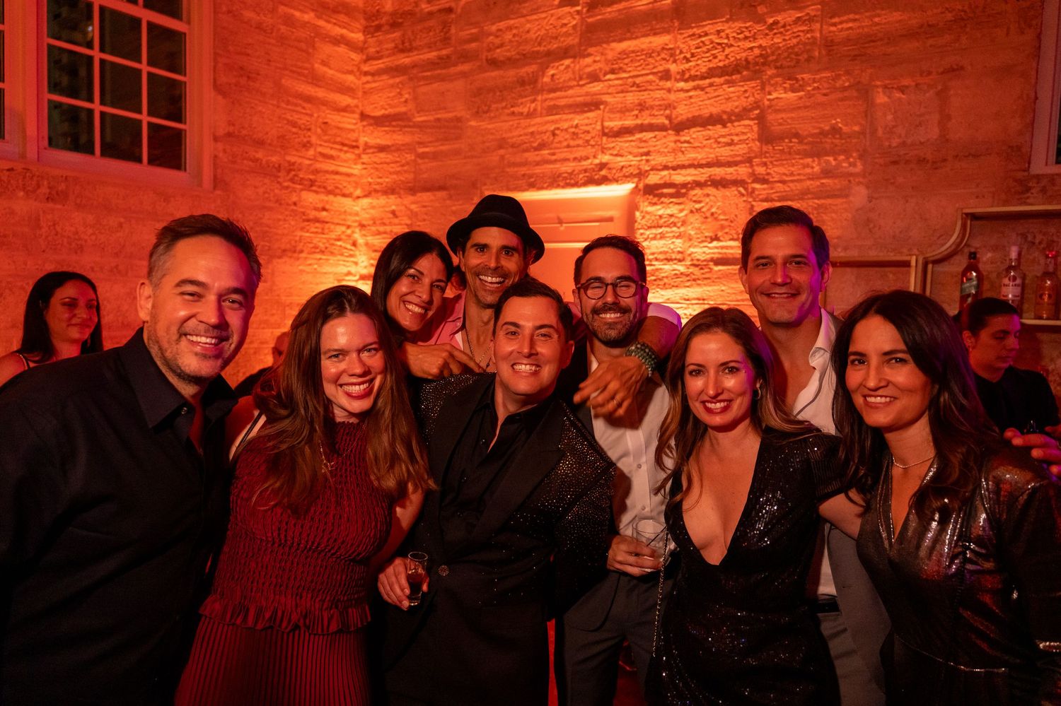 Group of friends smiling and posing together at a festive evening party with warm red ambient lighting.