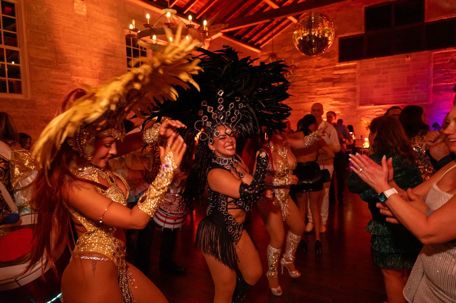 Samba dancers in vibrant carnival costumes with feathered headdresses perform at a festive indoor event.