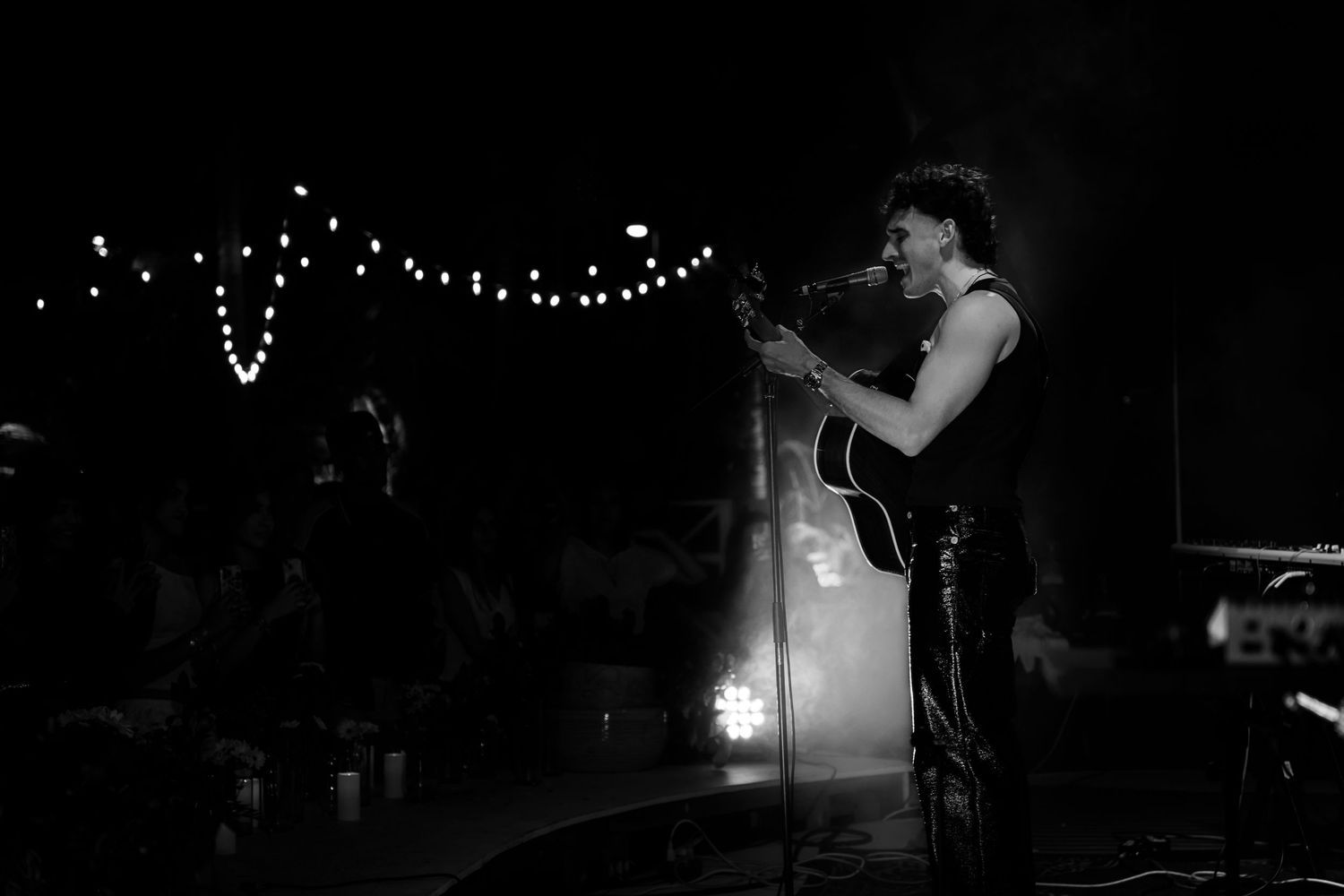 A performer sings passionately on stage in a dark venue with string lights glowing in the background.