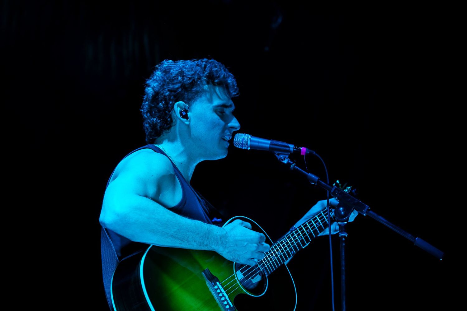 Musician performs on acoustic guitar in dramatic blue and green stage lighting during live concert performance.