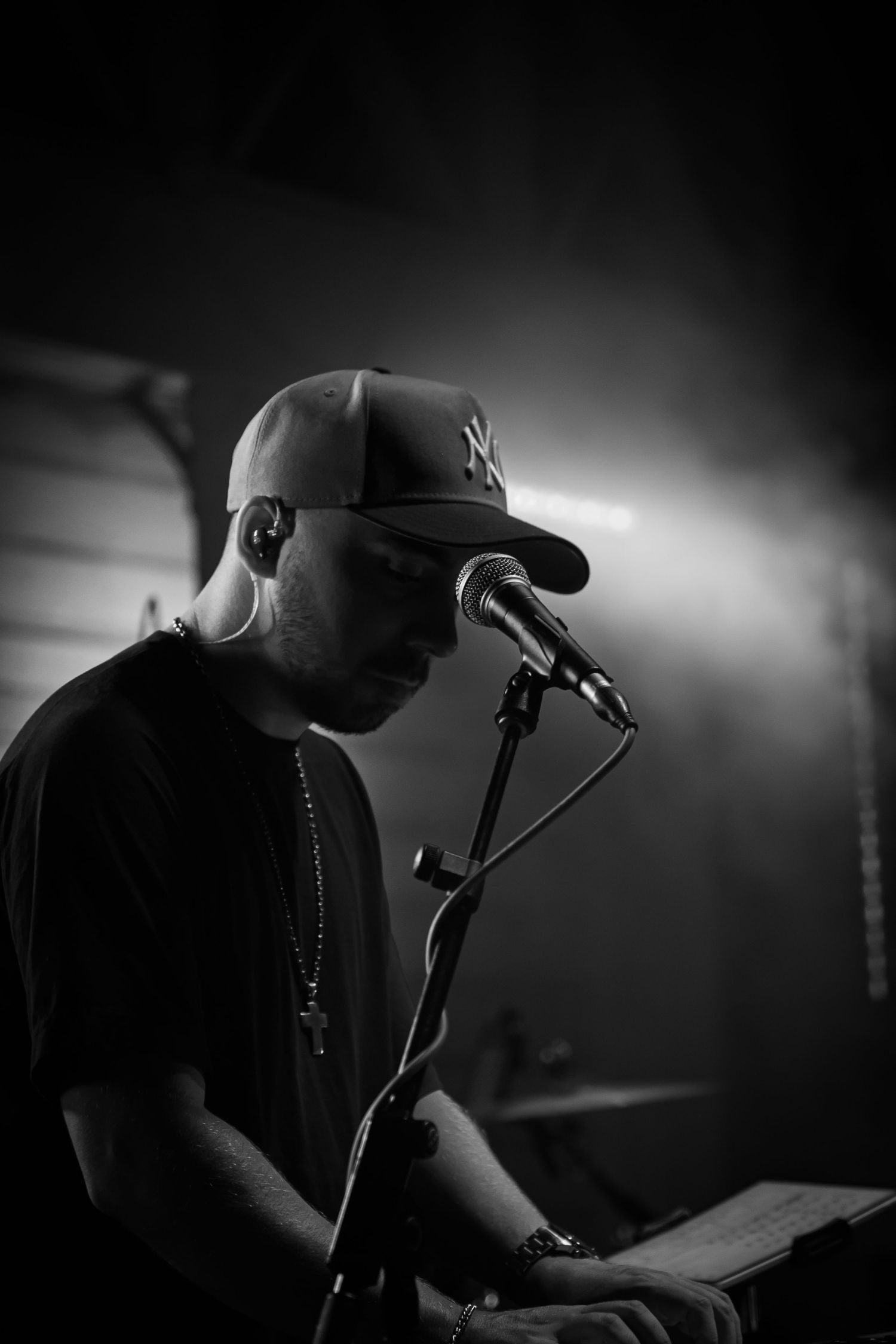 Black and white photo of a performer in a NY Yankees cap singing into a microphone during a moody stage performance.