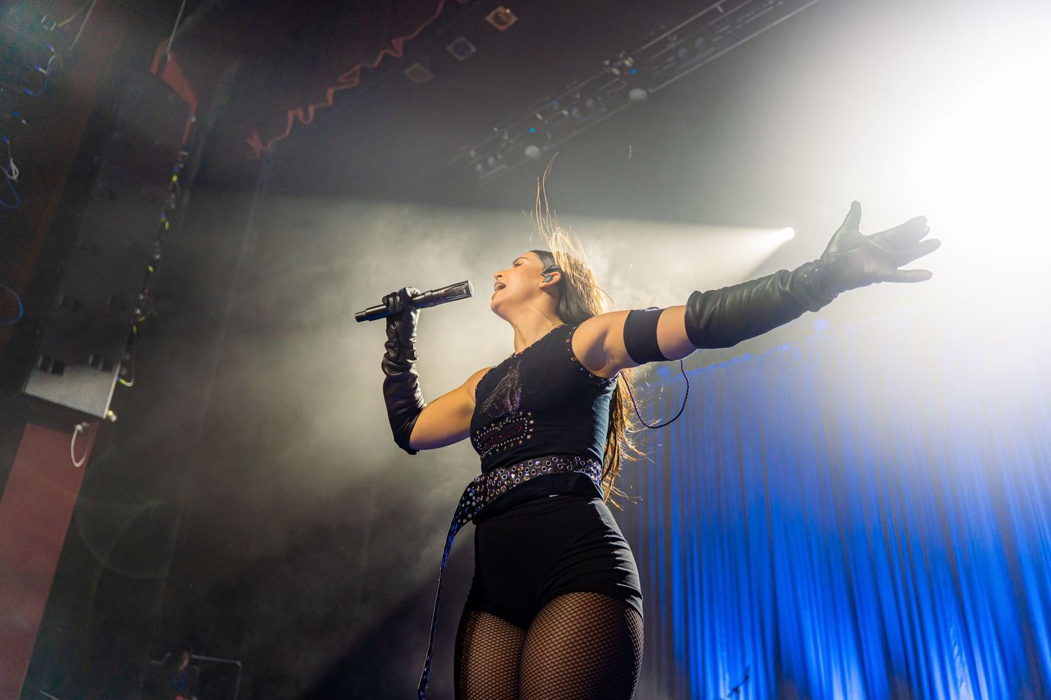 Nathy Peluso in black outfit sings passionately on stage with arms outstretched amid dramatic lighting and smoke effects.