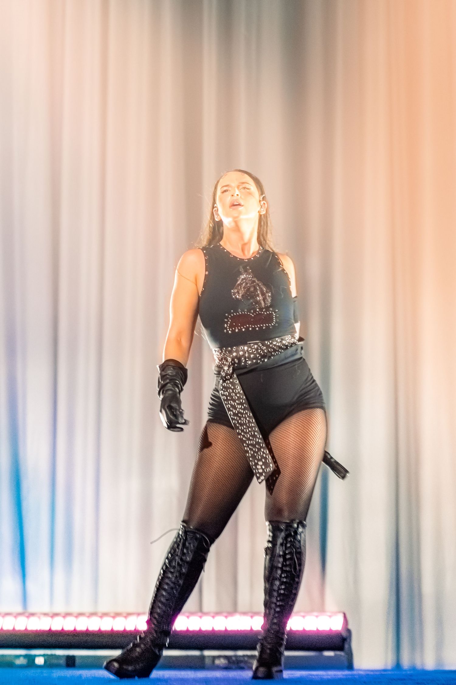 Female performer on stage in black bodysuit, chain belt, and knee-high boots, singing under warm stage lighting.