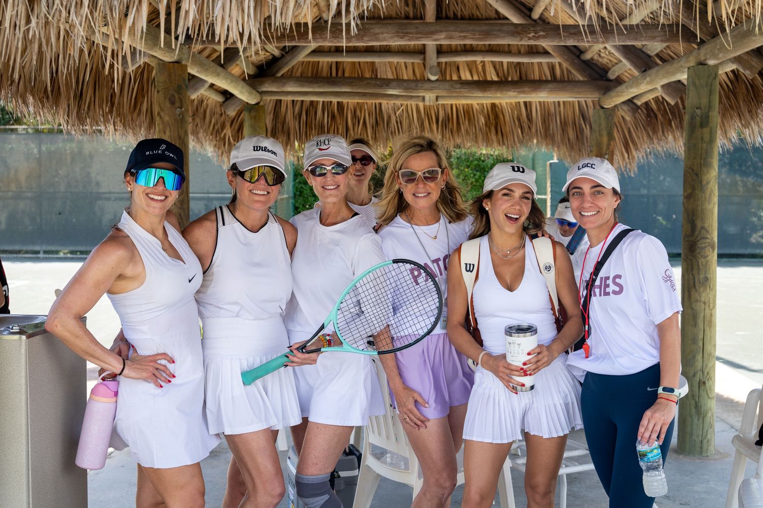 Seven women in white tennis outfits pose together under a thatched gazebo on a sunny day, one holding a tennis racket.