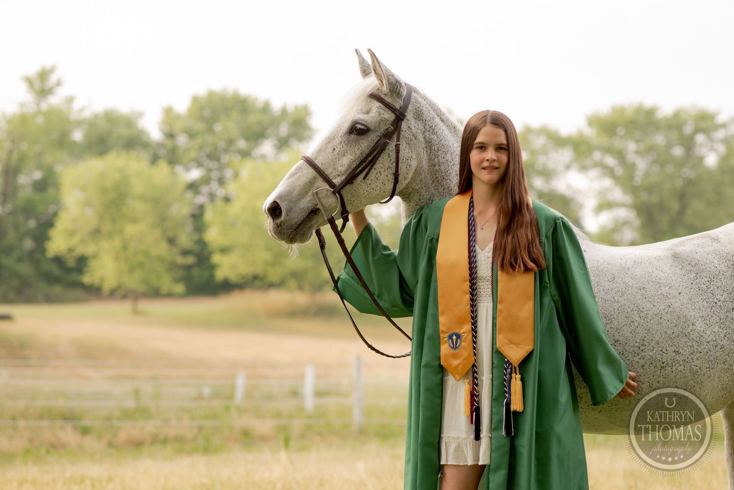 Graduate wearing green and gold regalia stands proudly next to a white horse in a field.