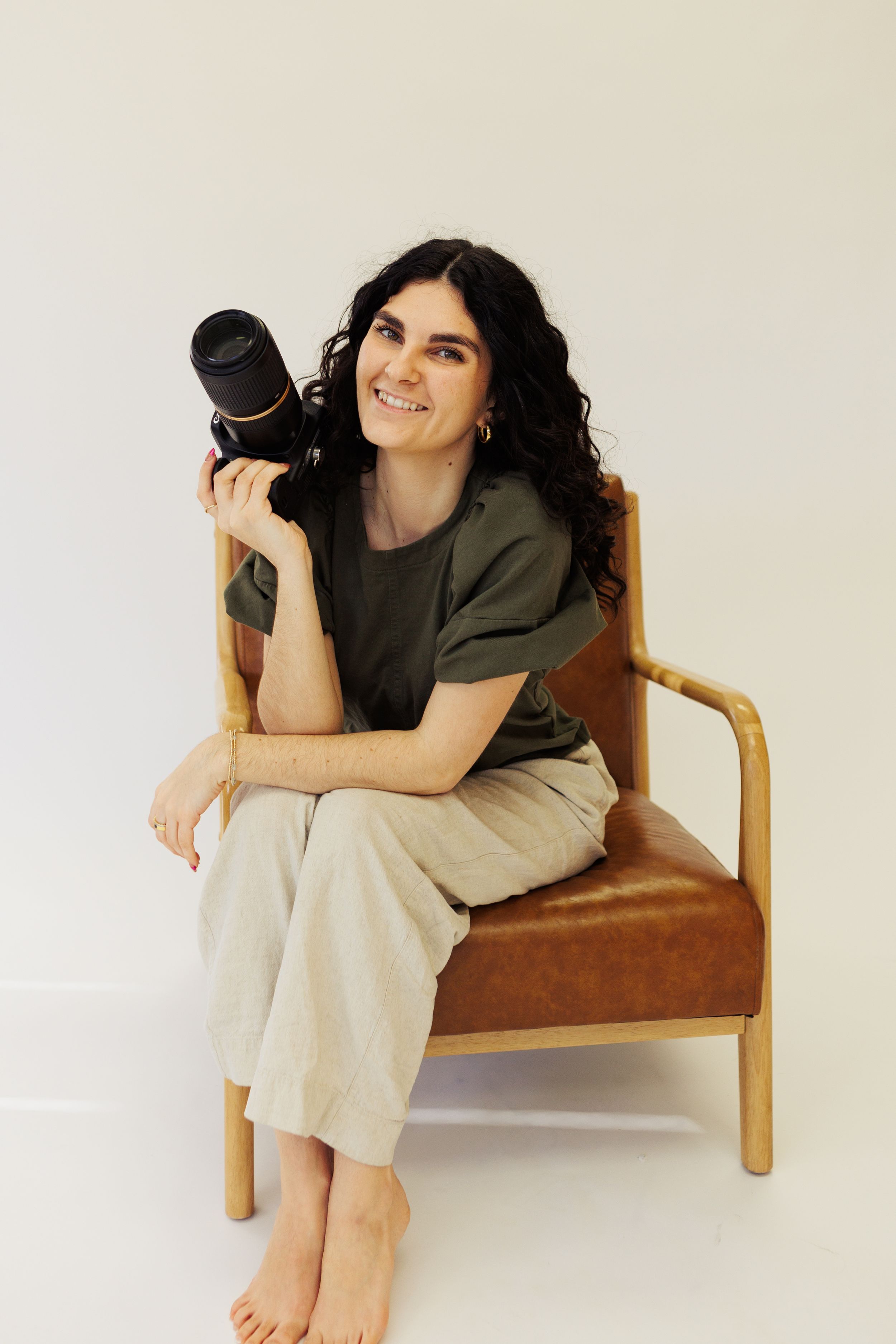 Photographer sitting barefoot in brown armchair holding camera lens while wearing olive shirt and beige pants.