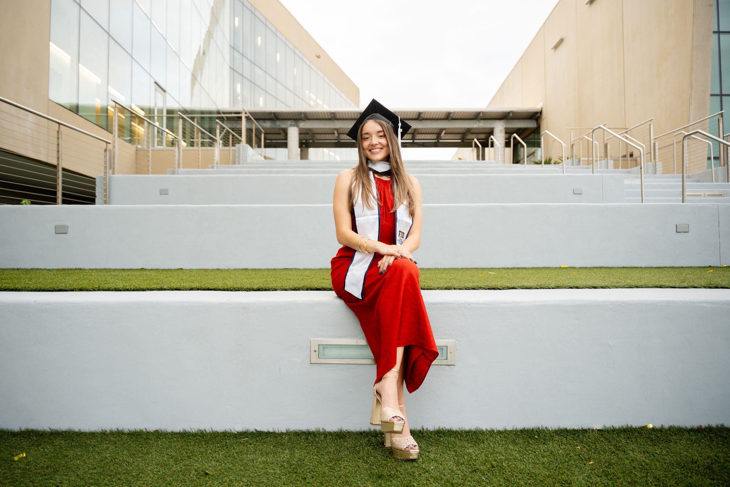Graduate in red and white regalia sitting on outdoor wall.