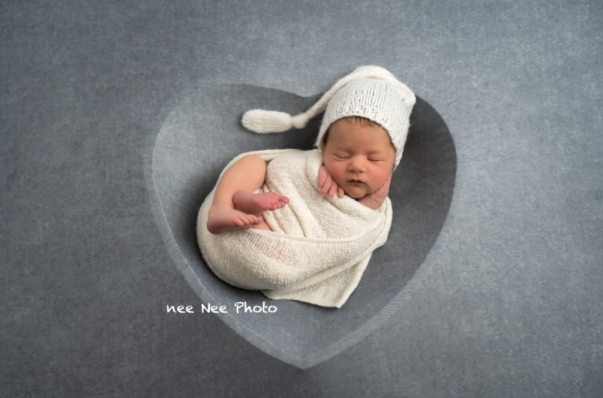 Newborn baby wrapped in white swaddle and knit hat, sleeping peacefully in a heart-shaped gray bowl. Embracing Life’s Beauty