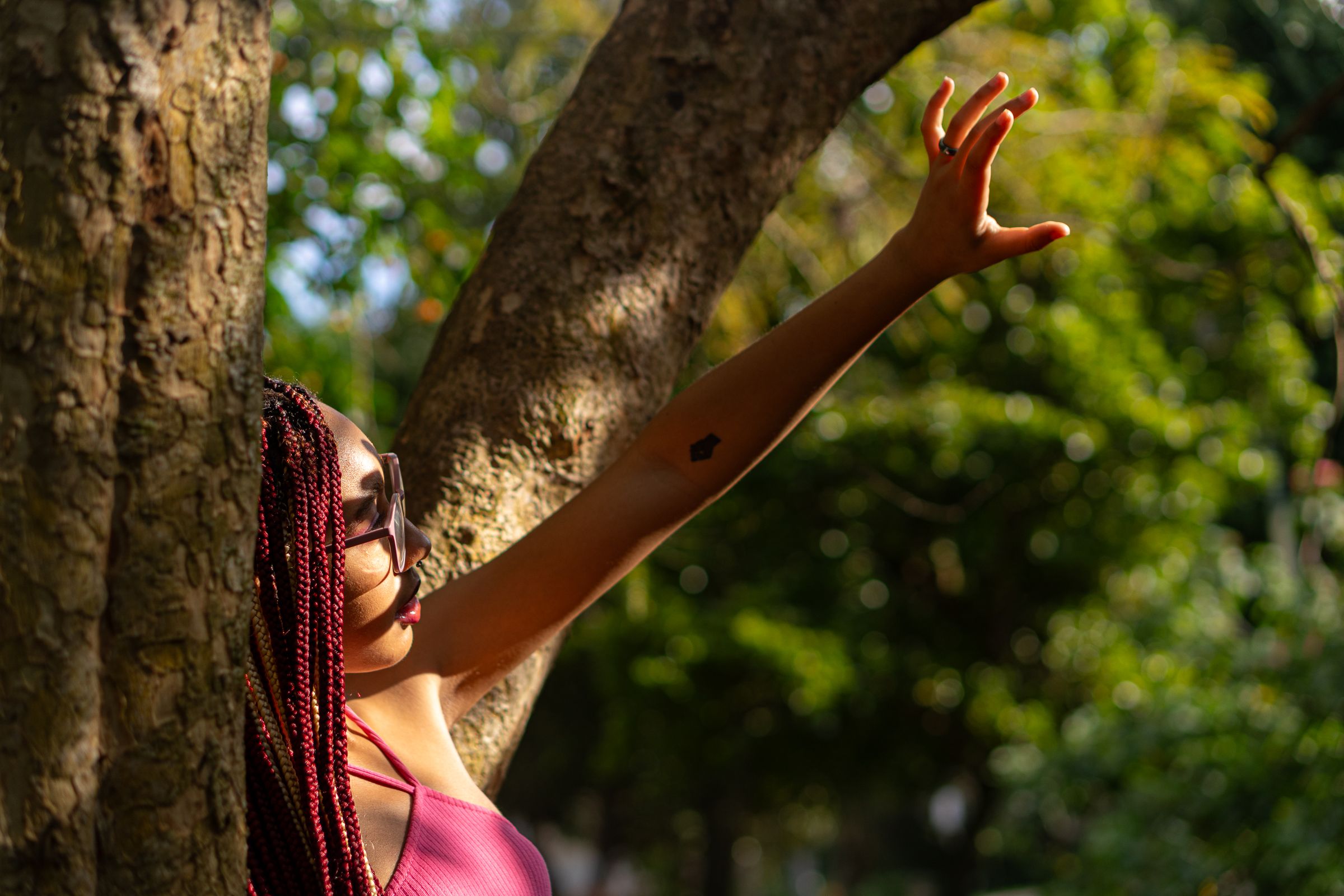 Woman with pink hair reaches up towards sunlight in nature setting.