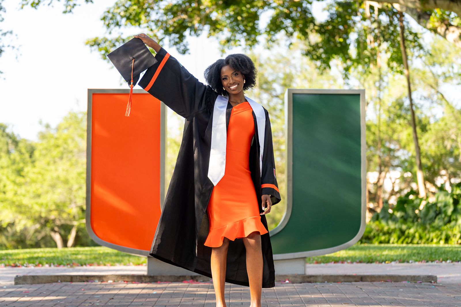 Graduate in black gown celebrating in front of university logo wall.