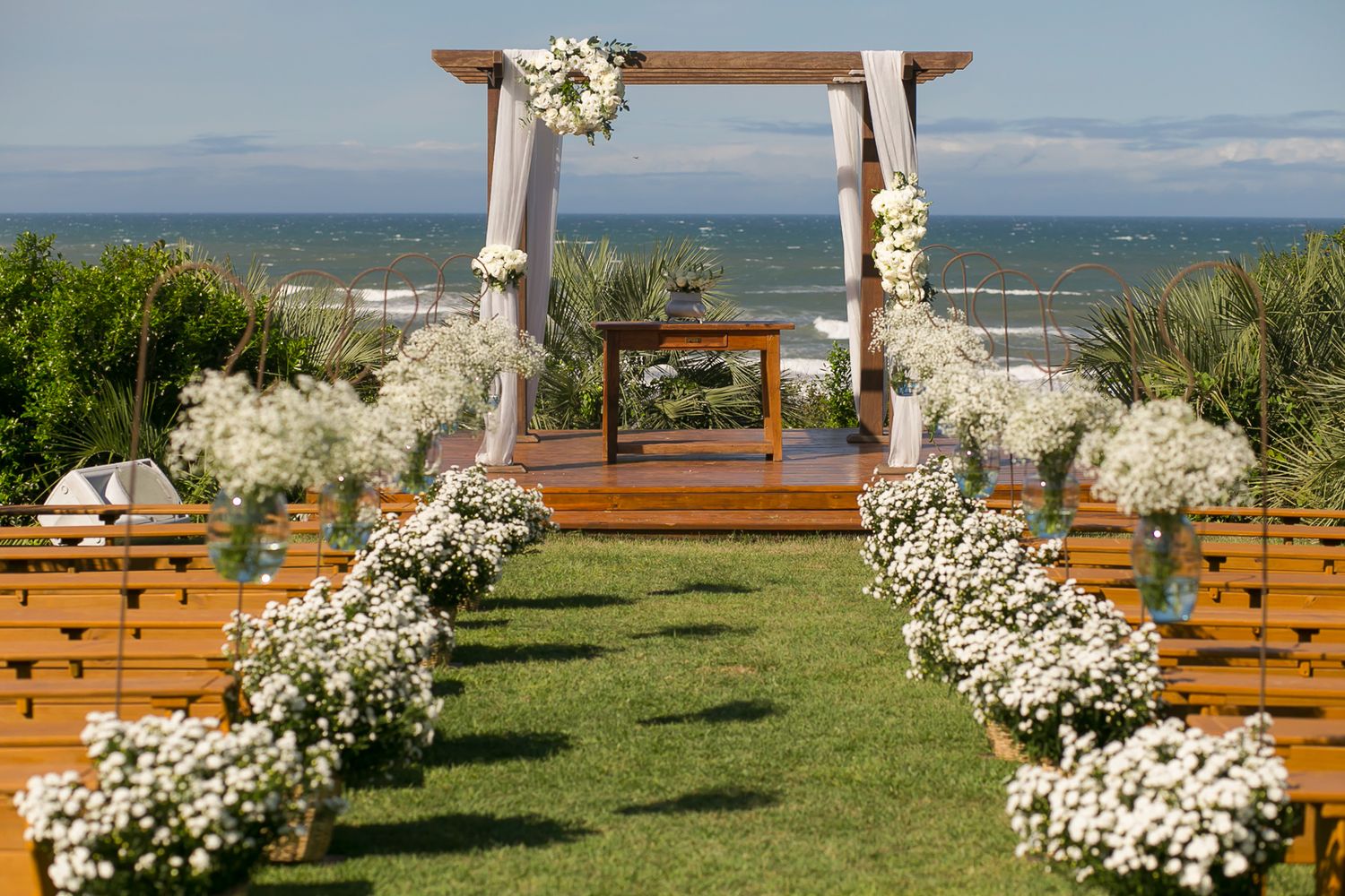 Outdoor wedding ceremony setup with white floral arch and wooden benches overlooking the ocean.