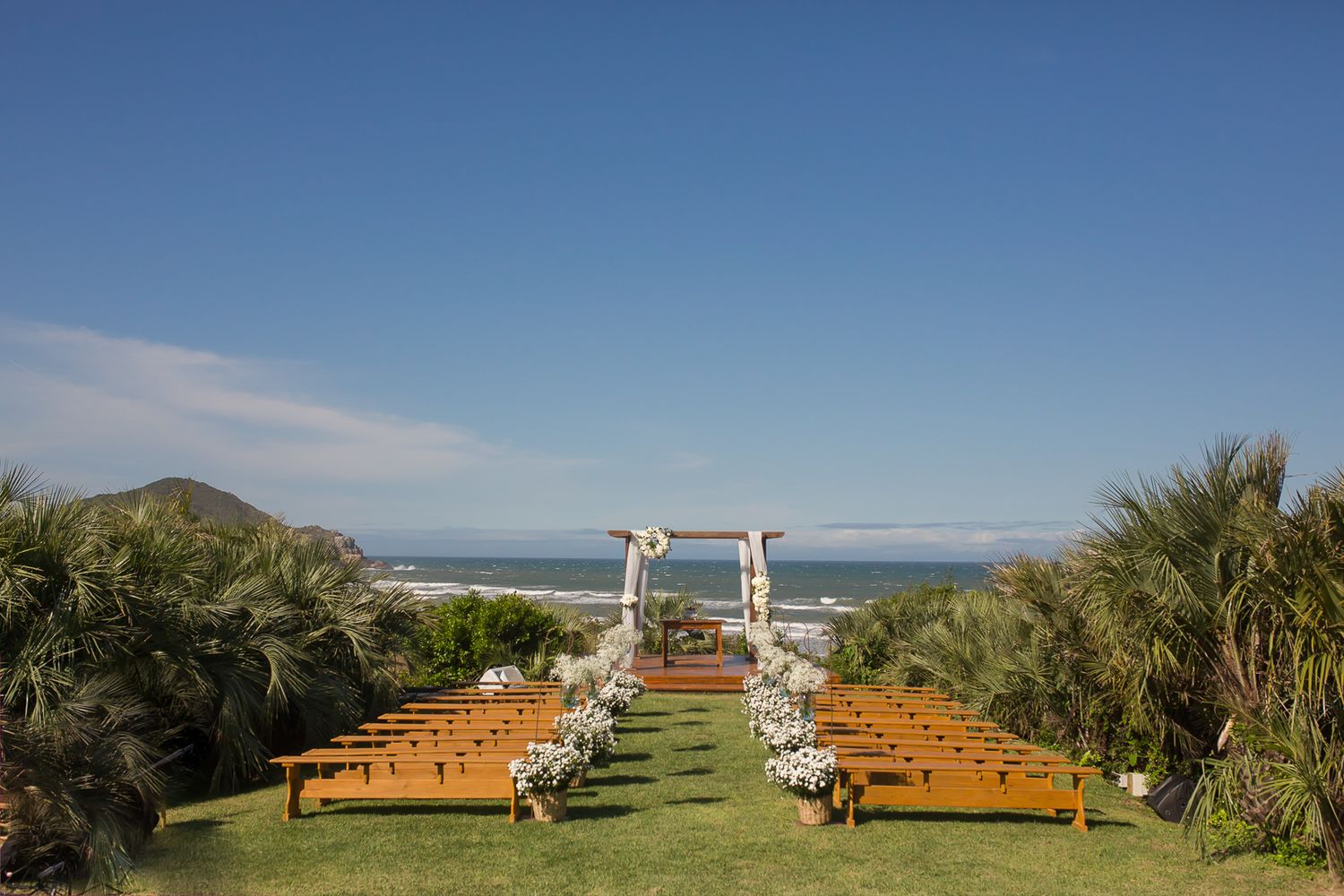 Coastal wedding venue setup with wooden benches and decorated arch against ocean backdrop.