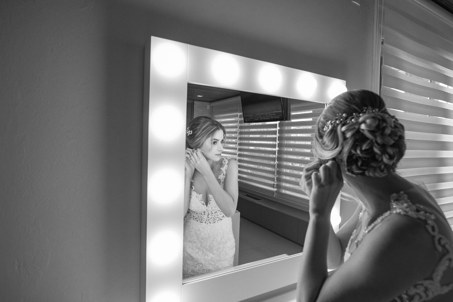 Black and white reflection of bride getting ready in illuminated makeup mirror.