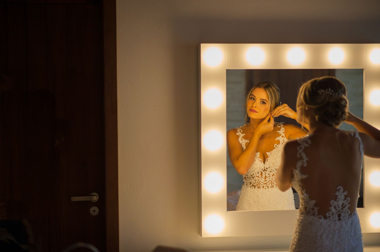 Bride preparing in front of illuminated vanity mirror in warm lighting.