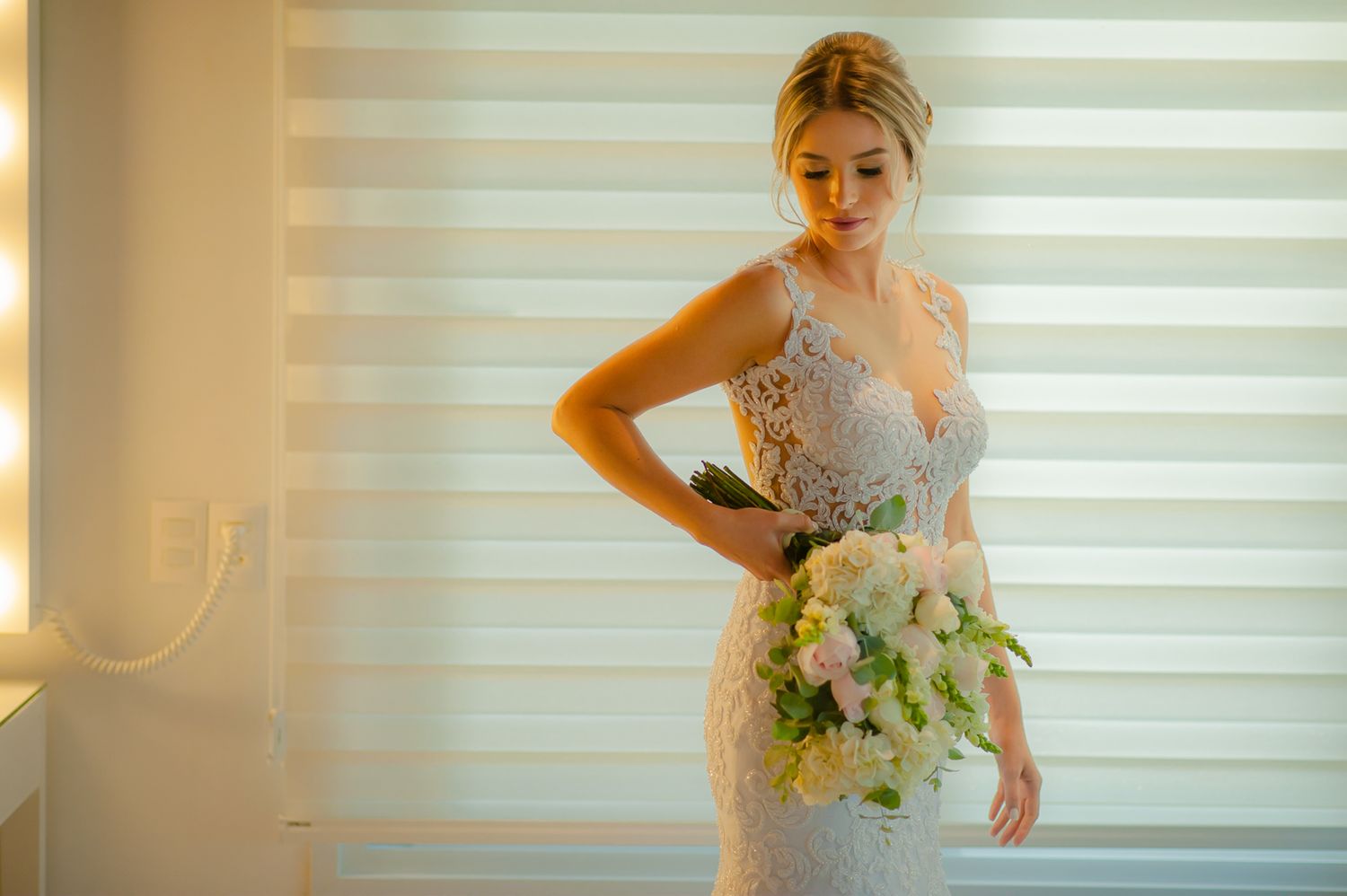 Elegant bride in lace dress poses with white bouquet against striped background.