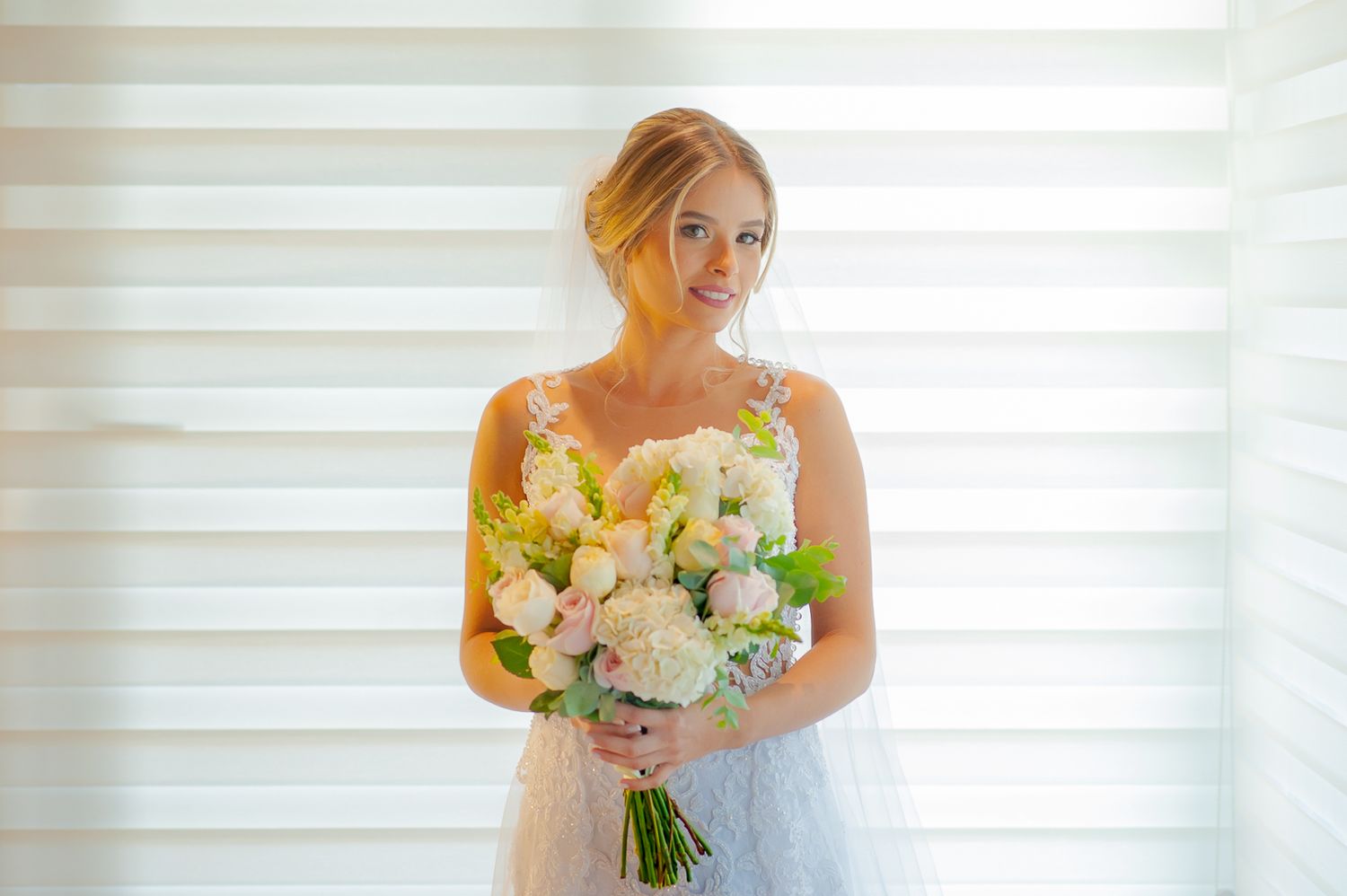 Radiant bride in lace dress holds fresh white and yellow bouquet against bright window.