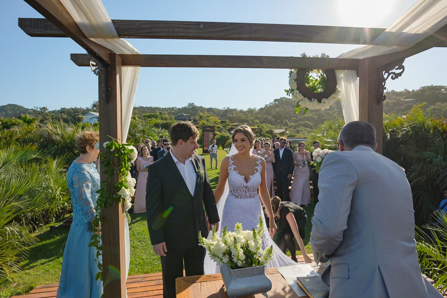 Outdoor wedding ceremony under a wooden pergola with tropical landscaping and mountains in the background.