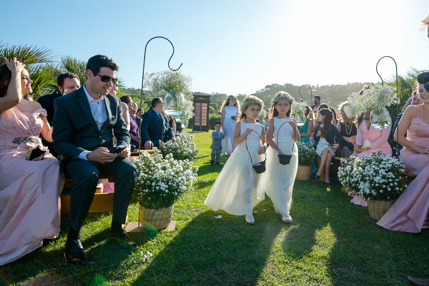 Young flower girls in white dresses walk down the aisle at an outdoor wedding ceremony.