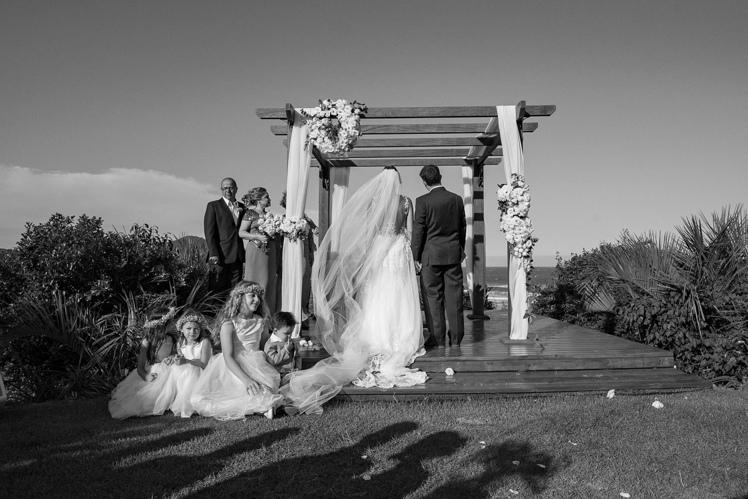 Black and white photo of wedding ceremony under rustic wooden arch with gathered guests.