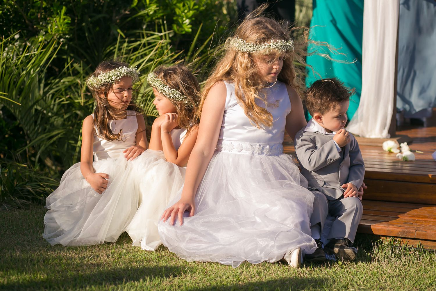 Flower girls in white dresses and young ring bearer sit together on wooden platform during ceremony.