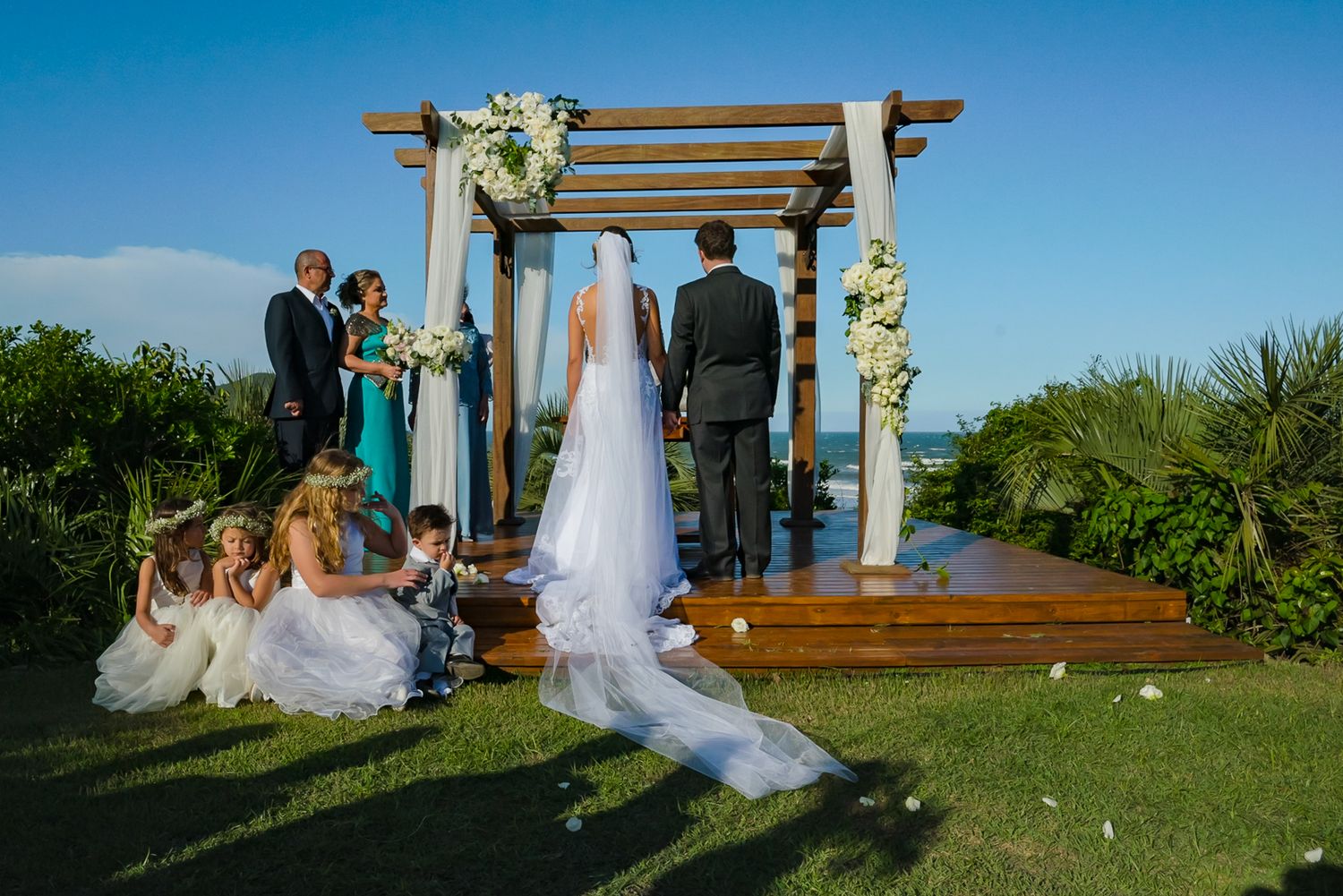A wedding ceremony takes place under a wooden pergola on a hillside overlooking the ocean.