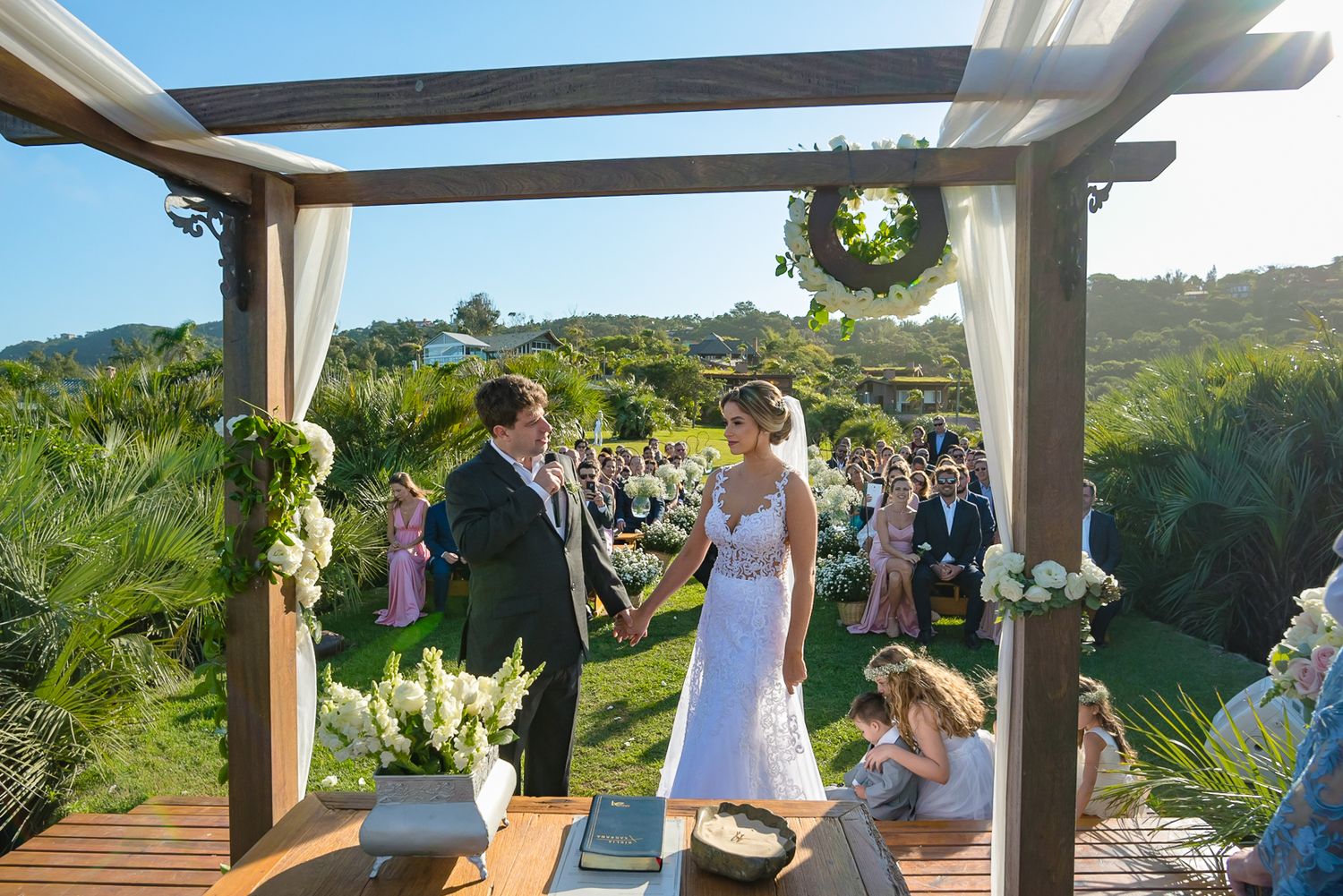 An outdoor wedding ceremony on a grassy hilltop with guests seated under a decorated wooden arbor.