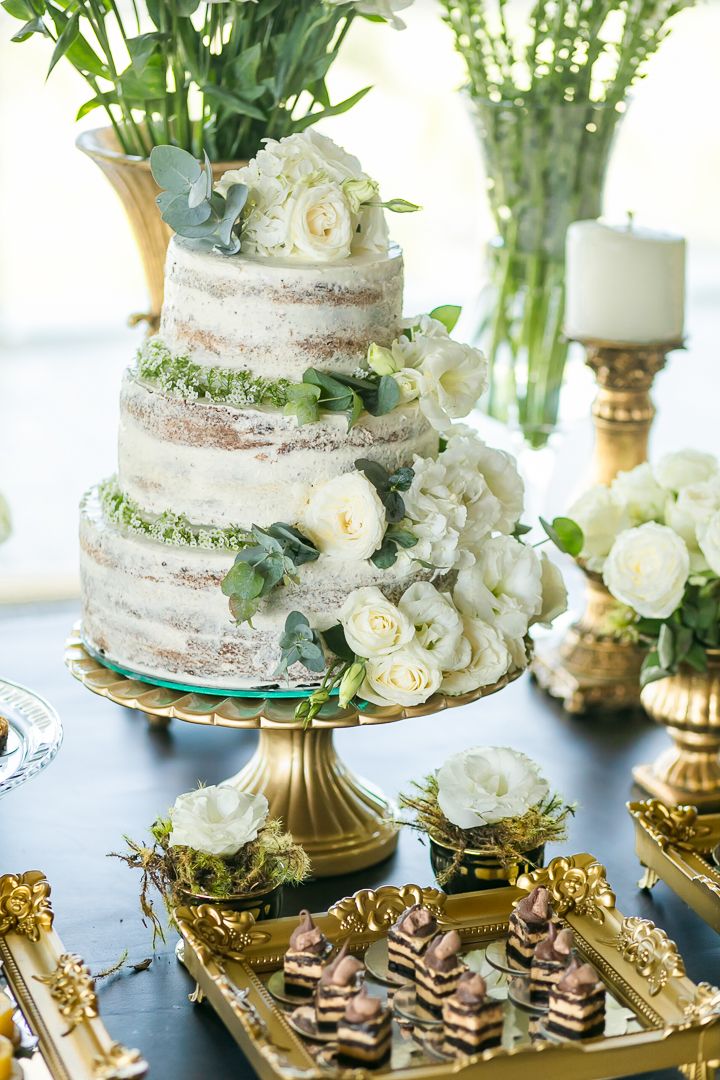 A naked wedding cake decorated with white roses and greenery on a gold stand.