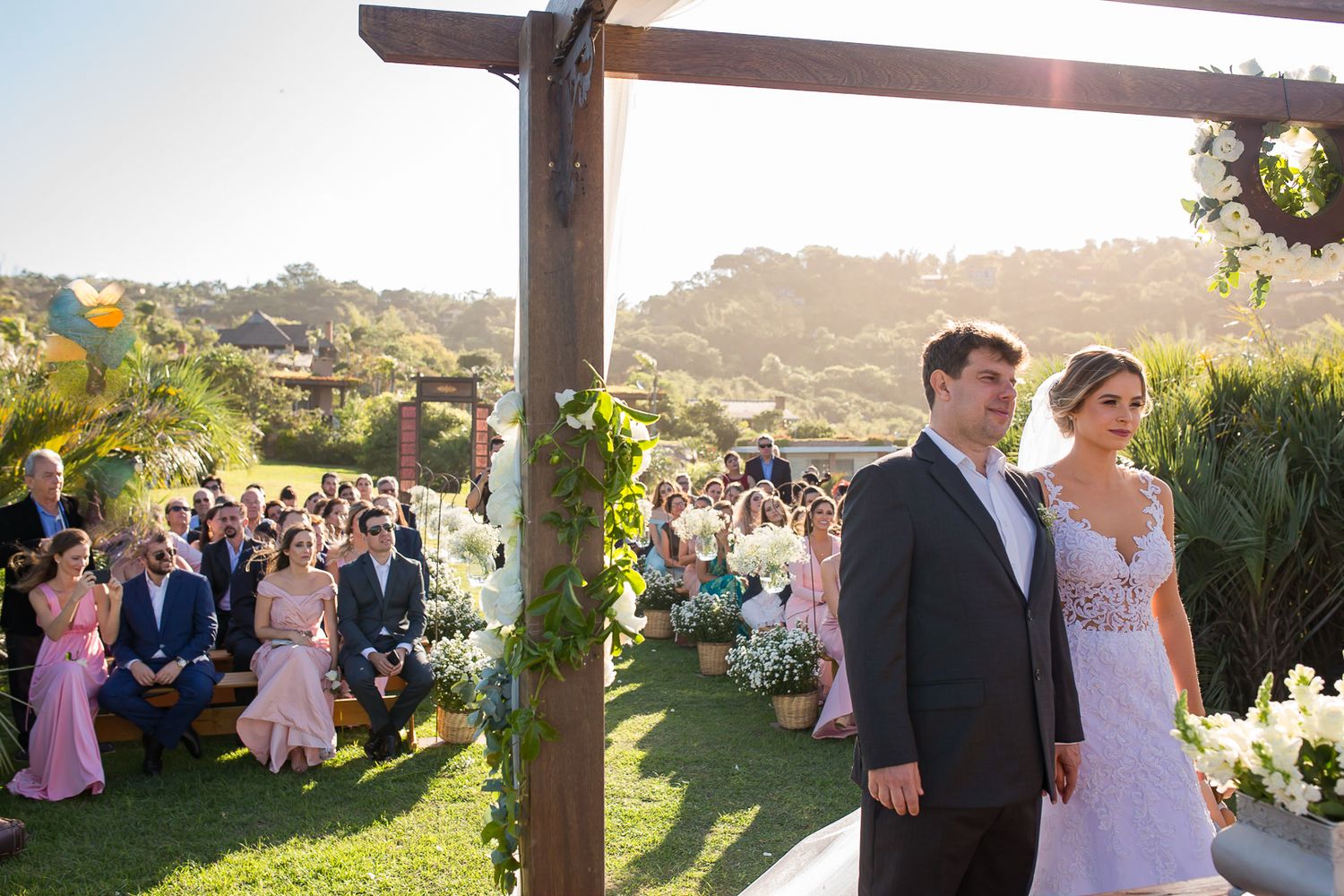 Wedding guests gather on a grassy hillside under a wooden pergola during a sunny afternoon ceremony.