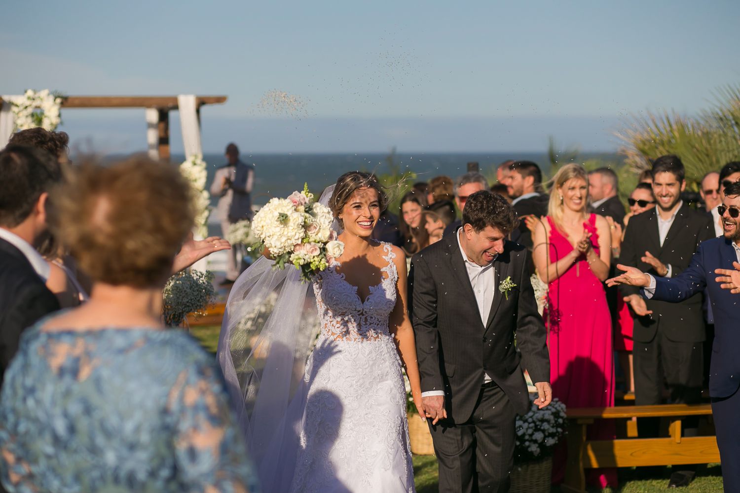 Wedding processional on a clifftop venue with ocean views and decorated wooden pergola.