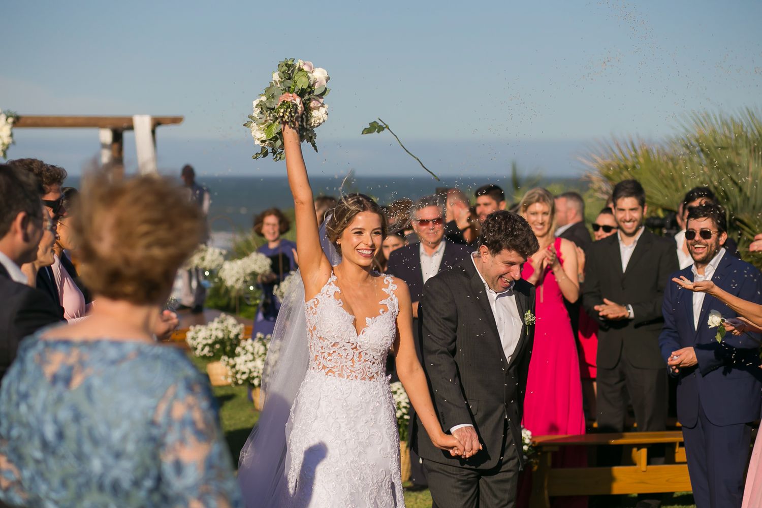 Joyful wedding celebration under blue skies with ocean backdrop and wooden ceremony arch.