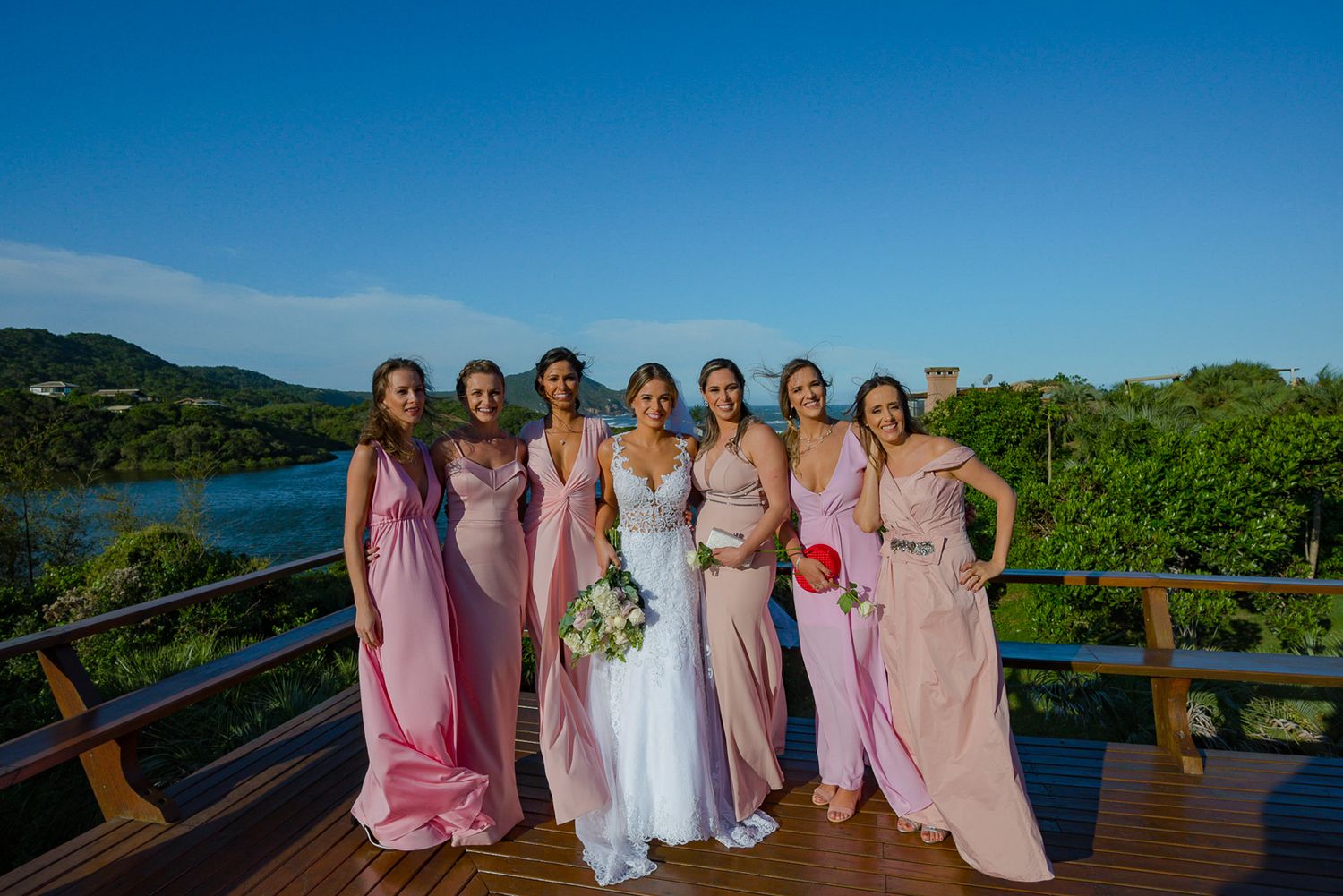 Wedding party in pink bridesmaid dresses posing on wooden deck with ocean view backdrop.